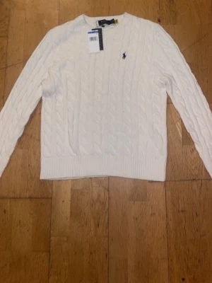 Vit kabelstickad tröja Polo Ralph Lauren - Kabelstickad vit tröja från Polo Ralph Lauren med rund halsringning och den klassiska broderade loggan på bröstet. Tröjan har långa ärmar och ribbade muddar vid ärmslut och nederkant. Perfekt för en clean och stilren look.