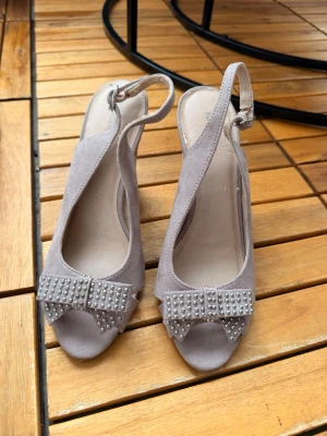 Ljuslila pumps med rosett och nitar - Säljer ett par ljuslila pumps med öppen tå och snygg rosettdetalj framtill dekorerad med silvriga nitar. Skorna har slingback-rem och klack, tillverkade i mockaimitation för en mjuk känsla. Perfekta för dig som vill sticka ut lite extra.