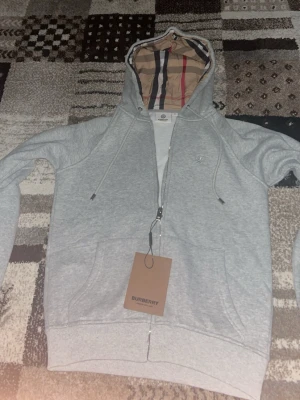 Grå zip hoodie från Burberry - Snygg grå hoodie från Burberry med dragkedja och klassiskt rutigt foder i huvan. Hoodien har dragsko i huvan, fickor framtill och ribbade muddar. Perfekt för en avslappnad men stilren look. Materialet är mjukt och bekvämt, troligen bomullsblandning.
