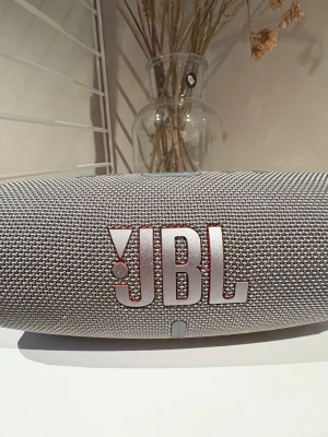 Grå JBL bärbar högtalare - Snygg grå bärbar högtalare från JBL med robust tygmaterial och rundad form. Perfekt för att ta med sig överallt och spela musik trådlöst. Stora JBL-loggan framtill och diskret rutmönster på tyget ger en modern look.
