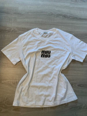 Vit Miu Miu t-shirt med logga - Säljer en vit t-shirt från Miu Miu med korta ärmar och rund hals. På bröstet finns en snygg Miu Miu-logga i svart med leopardmönster. T-shirten är i mjuk bomull och har en klassisk passform som funkar till allt.