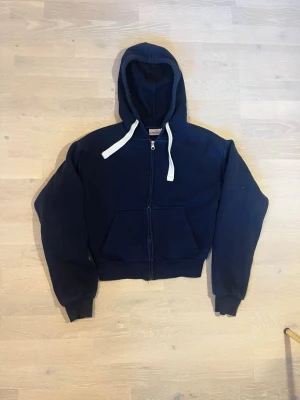 Mörkblå zip hoodie med huva gina tricot - En mörkblå hoodie med dragkedja framtill och stora fickor. Tröjan har en rejäl huva med vita snören och ribbade muddar vid ärmar och midja. Perfekt för en avslappnad och cool stil. Materialet känns mjukt och bekvämt.