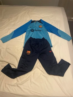 Barcelona träningsset Nike blå - Säljer ett Barcelona träningsset från Nike. Tröjan är ljusblå med klubbmärke och Spotify-logga, lång ärm och dragkedja vid halsen. Byxorna är mörkblå med klubbmärke och röd Nike-logga, smal passform och resår i midjan. Perfekt för fotbollsträning.
