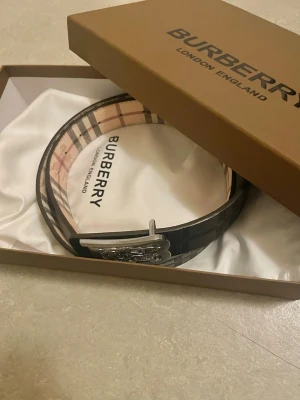 Randigt bälte från Burberry - Snyggt bälte från Burberry med klassiskt randigt mönster i beige, svart och rött. Bältet har ett stilrent silverfärgat spänne med Burberry-logga. Perfekt accessoar för att lyfta din outfit.