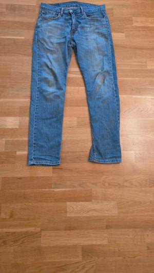 Blå Levis raka jeans med fem fickor - Klassiska blåa Levis jeans med rak passform och fem fickor. Jeansen har normal midja, dragkedja och knapp framtill samt bälteshällor. Materialet är denim i bomull och färgen är ljusblå med lätt slitning på låren och knäna.