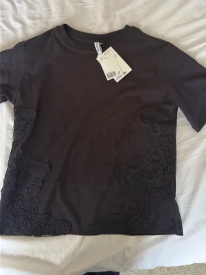 Svart t-shirt med spetsdetaljer - Snygg svart t-shirt med korta ärmar och dekorativa spetsdetaljer på sidorna. Tillverkad i mjuk bomull som känns skön mot huden. Perfekt för dig som vill ha en basic t-shirt med en twist.