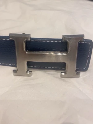 Blått skärp med silverfärgat H-spänne - Snyggt blått bälte med ett stort, silverfärgat H-format spänne. Bältet har vita kontrastsömmar längs kanterna och är gjort i skinn med en stilren och modern look. Perfekt accessoar för att lyfta din outfit.