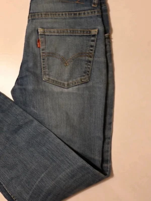 Blå Levi's raka jeans 152-164 - Säljer ett par klassiska blå Levi's jeans med raka ben och femficksmodell. Jeansen har en ljus tvätt och den ikoniska röda Levi's-lappen på bakfickan. Tillverkade i slitstark denim, perfekta för en avslappnad och tidlös stil.