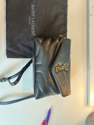 Svart axelväska från Saint Laurent - Säljer en svart axelväska från Saint Laurent i quiltat läder med guldigt YSL-emblem på framsidan. Väskan har justerbar axelrem och klassisk kuvertformad design. Perfekt för dig som vill ha en stilren och lyxig accessoar.
