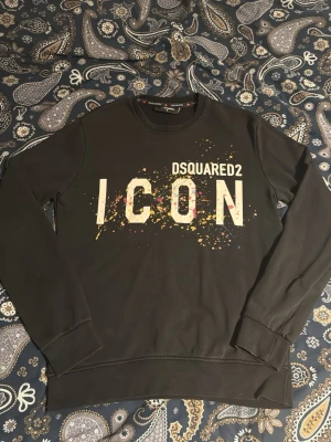 Svart Dsquared2 ICON sweatshirt - Svart sweatshirt från Dsquared2 med stor vit 'ICON'-text och färgstänk på bröstet. Rund halsringning och långa ärmar. Perfekt för dig som gillar streetwear och vill sticka ut med en trendig look.