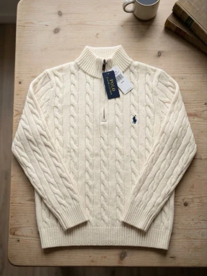 Vit kabelstickad tröja Polo Ralph Lauren - Kabelstickad tröja i offwhite från Polo Ralph Lauren med halv dragkedja och ribbad hög krage. Tröjan har långa ärmar, ribbade muddar och den klassiska blå logotypen broderad på bröstet. Perfekt för lager på lager och riktigt snygg till jeans.