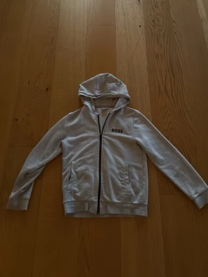 Ljusgrå hoodie med dragkedja från BOSS - Snygg ljusgrå hoodie från BOSS med dragkedja framtill och två fickor. Klassisk huva och ribbade muddar vid ärmslut och nederkant. Diskret BOSS-logga på bröstet. Perfekt för en chill och stilren look.