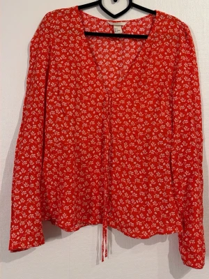 Röd blommig blus med knytning - Supersöt röd blus med små vita blommor och v-ringning. Långa ärmar och knytband framtill ger en avslappnad och trendig look. Perfekt att styla med jeans eller kjol för en fräsch vibe.