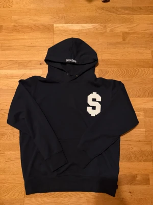 Mörkblå Supreme hoodie med $-tryck - Snygg mörkblå hoodie från Supreme med stort vitt $-tecken på bröstet. Perfekt för dig som gillar streetwear och vill sticka ut med en ikonisk detalj. Perfekt för höst som är runt hörnet. Aldrig använd