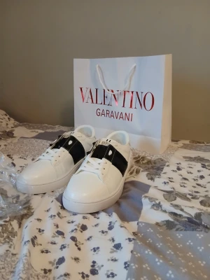 Vita sneakers med svart detalj - Snygga vita sneakers från Valentino med bred svart läderdetalj över sidan och vita snören. Klassisk låg modell med robust sula och diskreta nitar på hälen. Perfekta för dig som vill ha en clean men ändå unik look.