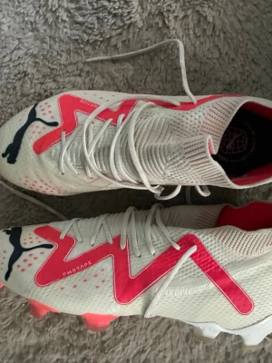 Vita och rosa Puma fotbollsskor - Säljer ett par snygga fotbollsskor från Puma i vitt med rosa och svarta detaljer. Skorna har snörning, mönstrad ovandel och dobbar för bra grepp på planen. Perfekta för dig som vill sticka ut på fotbollsplanen!