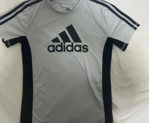 Vit Adidas t-shirt storlek 10/12 år - Snygg vit t-shirt från Adidas med svart logga på bröstet och svarta paneler längs sidorna. Klassiska svarta ränder på axlarna och korta ärmar. Perfekt för träning eller chill vardagsstil.