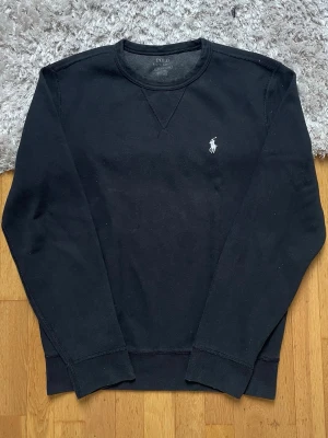 Svart sweatshirt från Polo Ralph Lauren - Klassisk svart sweatshirt från Polo Ralph Lauren med rund halsringning och den ikoniska vita logon broderad på bröstet. Tröjan har ribbade muddar vid ärmslut och nederkant, perfekt för en clean och stilren look.