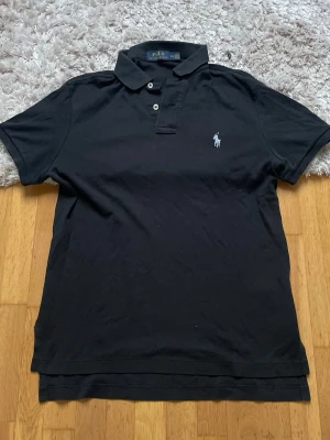 Svart pikétröja från Polo Ralph Lauren - Klassisk svart pikétröja från Polo Ralph Lauren med vit broderad logga på bröstet. Tröjan har korta ärmar, krage och två knappar vid halsen. Tillverkad i mjuk bomull för en bekväm känsla och stilren look.