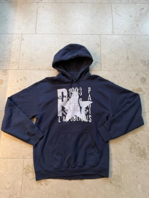 Mörkblå hoodie med tryck och huva - Säljer en mörkblå hoodie med stor vit stjärna och texttryck på bröstet. Hoodien har klassisk känguruficka, dragsko i huvan och ribbade muddar vid ärmar och nederkant. Perfekt för en avslappnad och cool stil.
