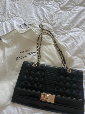 Svart quiltad handväska Donna Laura - Snygg svart handväska från Donna Laura med quiltat mönster, guldfärgade kedjehandtag och vridlås i guld. Väskan har en mix av glansigt och matt material som ger en lyxig känsla. Perfekt accessoar för att lyfta din outfit.