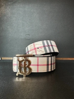 Burberry Bälte med Guld Spänne - Snyggt skärp i vitt med klassiskt rutigt mönster i svart och rött. Bältet har ett stort metallspänne i silverfärg med bokstavsdetalj. Perfekt accessoar för att lyfta din outfit.