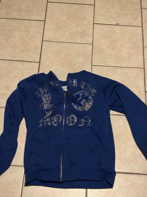Blå aim for the moon hoodie med tryck och dragkedja - Snygg blå hoodie med dragkedja framtill och huva. Framsidan har ett glittrigt tryck med texten 'AIM FOR THE MOON' och motiv av en tecknad figur och måne. Hoodien har ribbade muddar och fickor framtill. Perfekt för en chill och avslappnad stil.