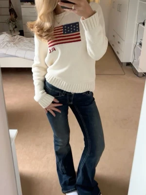 Polo Ralph Lauren flag sweater - Vit/cremefärgad stickad tröja från Polo Ralph Lauren med amerikansk flagga i blått och rött på bröstet samt RL-broderi. Tröjan har rund halsringning, ribbstickade muddar och är långärmad. Passar xs/s dam