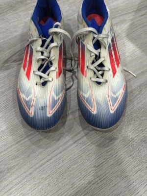 Adidas F50 League - Snygga fotbollsskor i vitt med blå och röda detaljer. Skorna har snörning och en sportig design med tunna ränder och kontrastfärgad insida. Perfekta för dig som vill sticka ut på planen och ha bra grepp och kontroll.