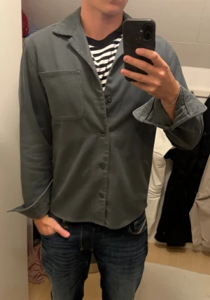 Grön overshirt  - Stilren grå overshirt från Dobber med klassisk krage, knappstängning och en bröstficka. Tillverkad i ett mjukt bomullstyg som ger en avslappnad look. Perfekt att bära över en t-shirt för en enkel och snygg outfit.
