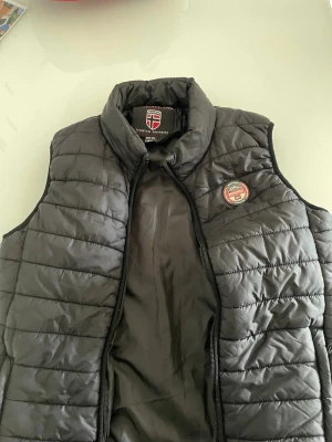 Svart dunväst från Geographical Norway - Snygg svart dunväst från Geographical Norway med quiltad design och hög krage. Västen har dragkedja framtill och ett runt märke på bröstet. Perfekt för lager-på-lager och håller dig varm under kyliga dagar.