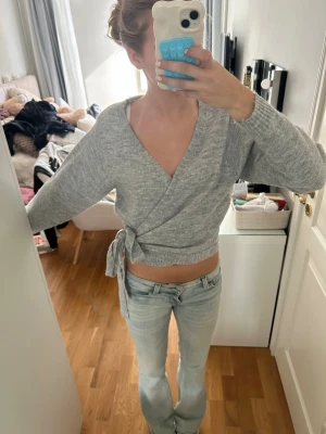 Grå omlottstickad tröja - Säljer en ljusgrå stickad omlottopp med knytband i sidan. Tröjan har lång ärm, djup v-ringning och ribbade muddar vid ärmslut och nederkant. Perfekt för lager på lager och enkel att styla till jeans eller kjol.