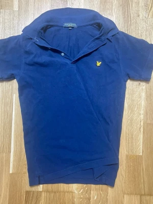 Mörkblå pikétröja från Lyle & Scott - Snygg mörkblå pikétröja från Lyle & Scott med klassisk krage och två knappar. Tröjan har det ikoniska gula örnemblemet broderat på bröstet. Tillverkad i mjukt bomullsmaterial som andas bra. Perfekt för en avslappnad och stilren look. Litet hål vid nacken men annars är allt okej