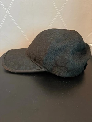 Svart keps med ton-i-ton GG-mönster - Snygg svart keps med ton-i-ton GG-mönster och böjd skärm. Kepsen har justerbart spänne bak i läder och är tillverkad i ett strukturerat tyg. Perfekt accessoar för en trendig streetwear-look.