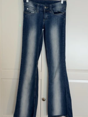 Blå bootcut jeans med slitningar - Snygga blå bootcut jeans med låg midja och med ljusa slitningar längs benen och klassisk femficksdesign. Jeansen har normal midja och utsvängda ben, vilket ger en retroinspirerad look. Tillverkade i denim med stretch för extra komfort. 📍STORLEKEN är w25 och L32📍
