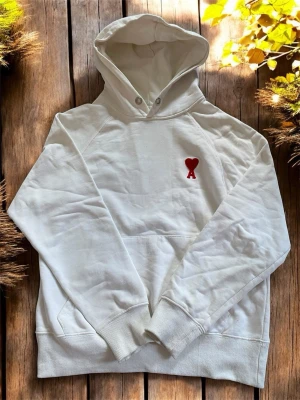 Vit hoodie med röd hjärtlogga - Snygg vit hoodie med huva och dragsko. På bröstet finns en liten röd hjärtlogga med bokstaven A. Hoodien har ribbade muddar vid ärmar och midja, samt en klassisk känguruficka framtill. Perfekt för en chill och stilren look.