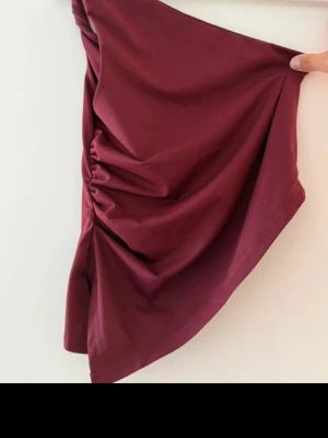 Vinröd oneshoulder tubtopp - Trendig vinröd tubtopp med oneshoulder-design och snygg drapering på sidan. Toppen har en asymmetrisk form och är gjord i ett mjukt, stretchigt material som sitter skönt mot kroppen. Perfekt för dig som vill ha en unik och stilren topp.