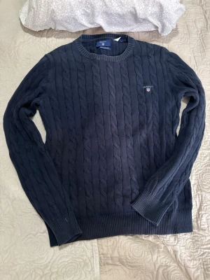 Mörkblå kabelstickad tröja GANT - Kabelstickad mörkblå tröja från GANT med rund halsringning och ribbade muddar. Tröjan har GANT-logga och sköld broderad på bröstet. Klassisk och tidlös design som passar perfekt till jeans eller chinos.