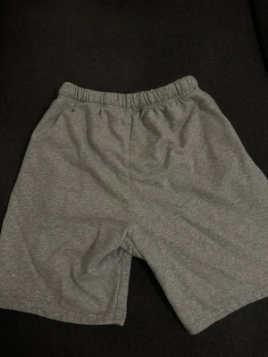 Grå sweatshorts från Essentials - Grå sweatshorts från Essentials med tryckt logga på vänster ben och elastisk midja med dragsko. Tillverkade i mjukt sweatshirtmaterial som ger en avslappnad och bekväm känsla. Perfekta för chill dagar eller streetwear-stil.