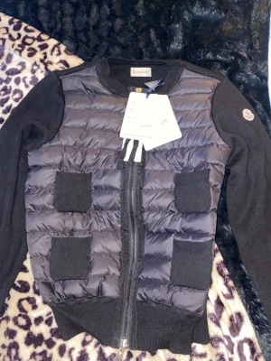 Svart cardiganjacka från Moncler - Snygg svart cardiganjacka från Moncler med quiltad framsida och stickade ärmar. Fyra framfickor i stickat material och dragkedja framtill. Moncler-logga på ärmen. Perfekt för lager på lager och streetstyle.
