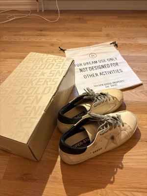 Golden Goose Superstar sneakers vita/svarta/gråa - Golden Goose Superstar sneakers i vitt läder med grå stjärndetalj på sidan och svarta hälpartier. Storlek 42 och i nyskick.