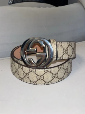 Beige GG-bälte med silverdetalj - Snyggt beige bälte  med det klassiska GG-mönstret och en stor, rund silverspänne med GG-logga. Bältet har en lyxig känsla och passar perfekt till jeans eller byxor för att lyfta din outfit.
