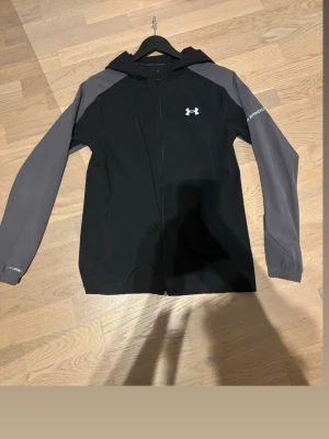 Svart och grå vindjacka Under Armour - Snygg svart vindjacka från Under Armour med grå ärmar och huva. Jackan har dragkedja framtill, logga på bröstet och är tillverkad i ett lätt och sportigt material. Perfekt för blåsiga dagar och träning utomhus. Modellen finns inte att köpa längre detta gör att jackan blir mer unik!