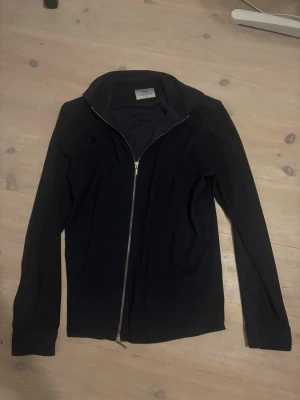 Ciszere tröja - Stilren svart ziptröja med hög krage och hel dragkedja framtill. Tröjan har långa ärmar och knappdetaljer vid ärmsluten. Perfekt för en clean och enkel look.