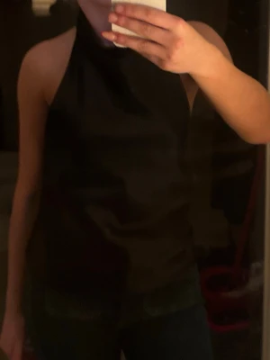 Svart halterneck topp - Säljer en svart halterneck topp med hög hals och ärmlös design. Toppen har en clean och stilren look, perfekt att styla med jeans eller kjol. Materialet ser ut att vara mjukt och följsamt, troligen polyester eller bomull.