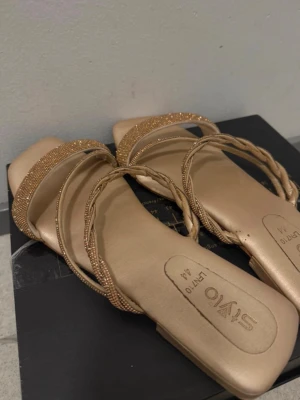 Guldiga sandaler från SYVO - Snygga guldiga sandaler från SYVO med fyrkantig tå och glittrande remmar. Remmarna är dekorerade med små stenar och en av dem är tvinnad för extra detalj. Perfekta för att lyfta din outfit med lite extra bling. Jag använder själv 43 men har svårt för att gå i sån sandaler tyvärr. Skulle säga dom mer är en 42 