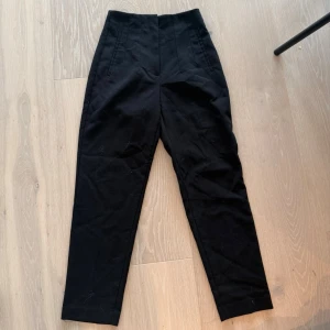 Svarta höga kostymbyxor Zara - Snygga svarta kostymbyxor med hög midja och avsmalnande ben. Byxorna har pressveck och diskreta fickor framtill. Perfekta för en clean och stilren look. Materialet ser ut att vara en blandning av bomull och polyester.
