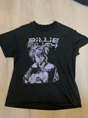Svart Billie Eilish t-shirt H&M - Cool svart t-shirt från H&M med stort Billie Eilish-tryck framtill i vitt och grått. Oversized passform och klassisk rund hals. Perfekt för dig som gillar musik och streetstyle.