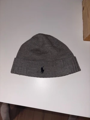 Grå mössa från Polo Ralph Lauren - Säljer en stilren grå mössa från Polo Ralph Lauren med klassisk ribbstickning och uppvikt kant. Mössan har den ikoniska mörkblå loggan broderad framtill och är tillverkad i mjukt material som håller dig varm under kalla dagar.