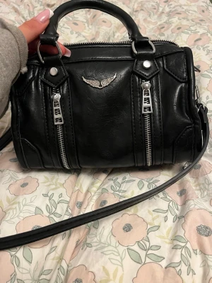 Zadig väska  - Cool svart handväska i läderlook med två silverfärgade dragkedjor framtill och ett snyggt emblem med vingar. Väskan har både handtag och en lång axelrem, perfekt för att bära på olika sätt. Silverfärgade metalldetaljer ger en edgy känsla.